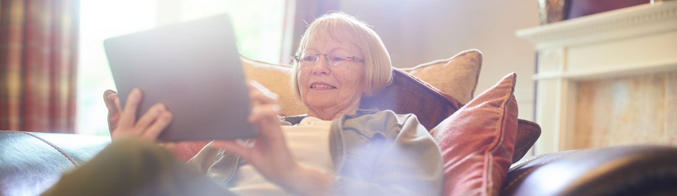 A woman sits on her couch smiling and using her electronic tablet.