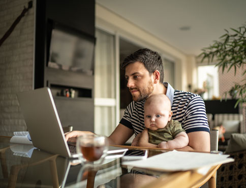 A dad holds his baby boy while sitting at the dining room table and working on his laptop.