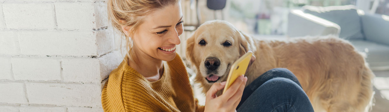 A woman sits on the floor smiling and looking at her cellphone with her Golden Retriever dog nearby.