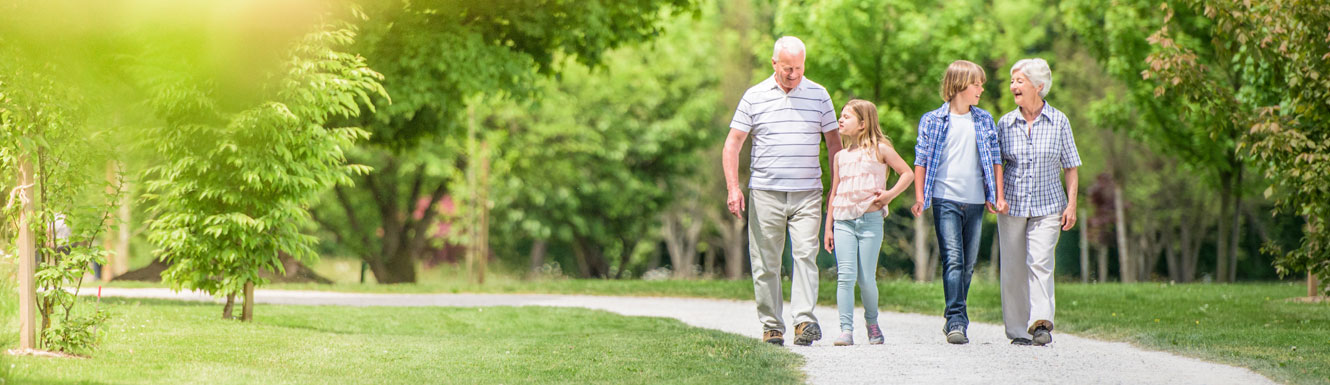 Two grandparents are walking with their young grandchildren on a path surrounded by grass and trees.