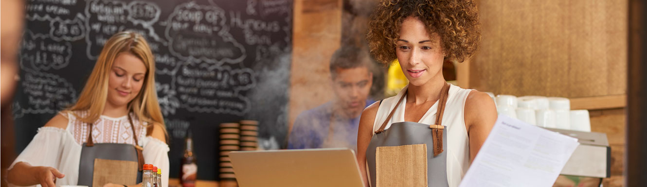 A small business owner young woman is using a laptop and holding paperwork in a coffee shop.