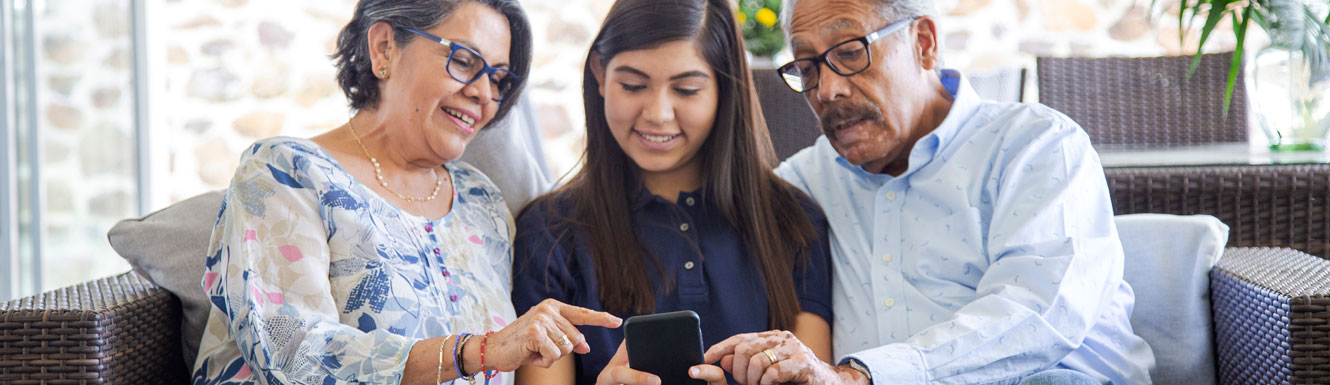 Two grandparents are smiling and showing their teenage granddaughter something on her cellphone.