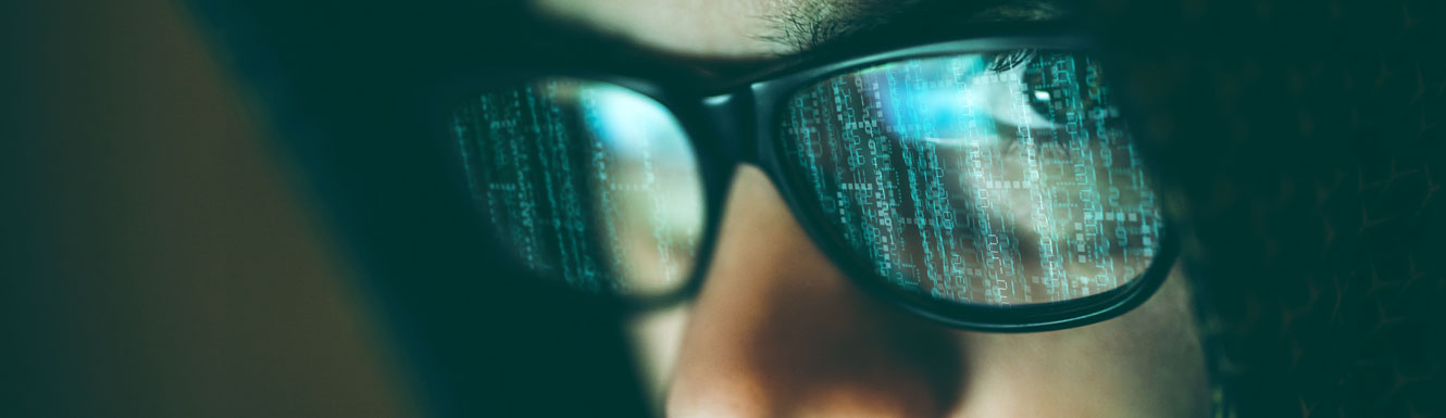 A closeup of a computer hacker with a computer screen reflecting in his eye glasses.
