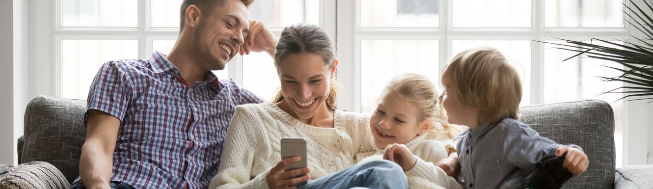 A happy young family is sitting on the couch and smiling while the mother uses her cellphone.