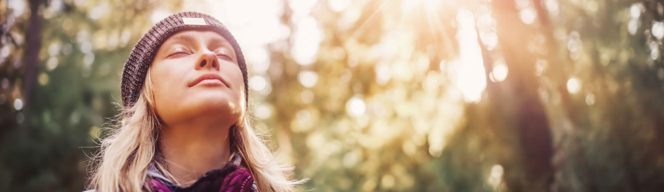 A woman with her eyes closed is in the forest and looking toward the sky.