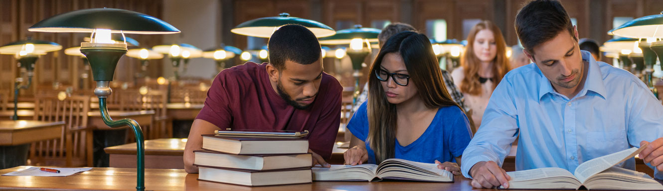 Three young people are sitting at a table with lamps reading in a large wood paneled library.