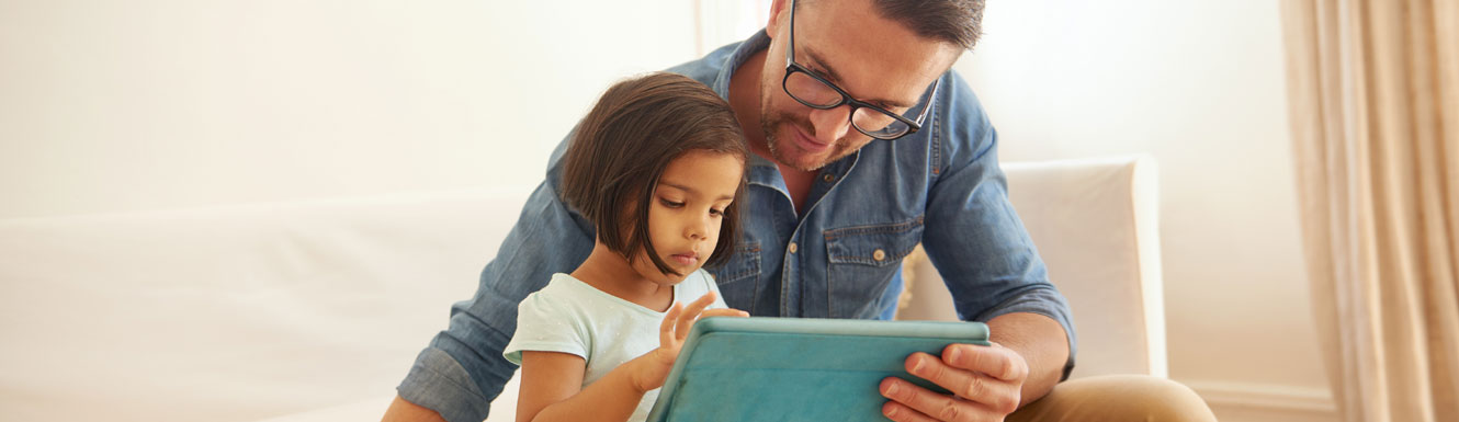 A father and his young daughter are sitting on the couch using an electronic tablet.