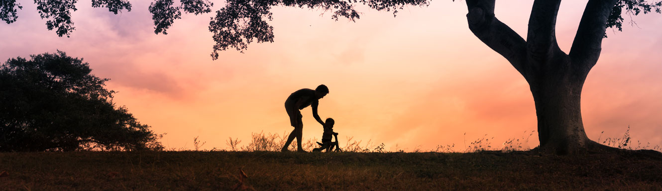 A father is pushing his toddler on a tricycle with a beautiful sunset in the background.