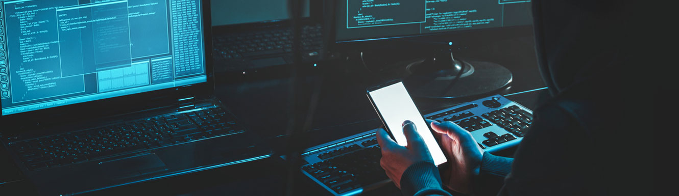 An individual in a hoody is shown in a dark room with a cell phone, sitting in front of numerous computer screens.