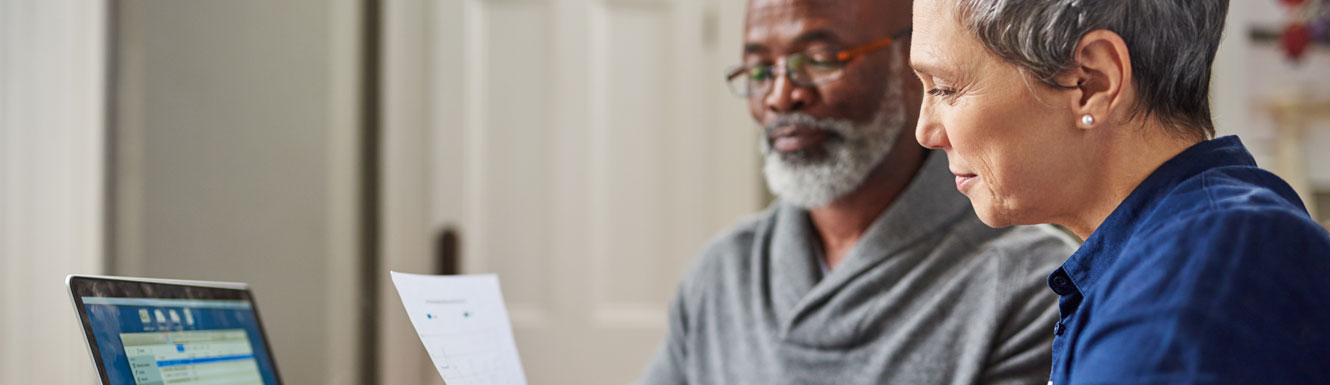 An older couple are sitting looking at paperwork and a computer.