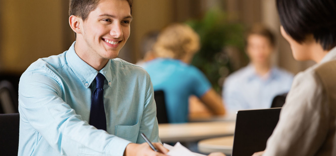 A smiling young man is at an interview with other people pictured in the background.