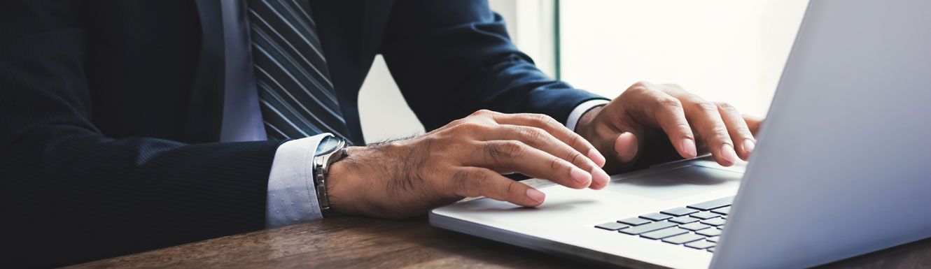 A business man in a dark suit with a striped tie is at his desk using a laptop.