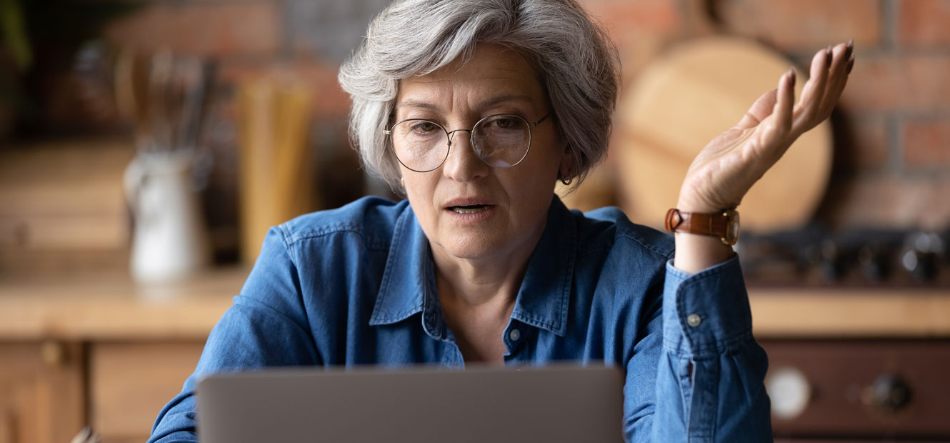 A mature woman in glasses is pictured looking at a laptop with a puzzled look on her face.