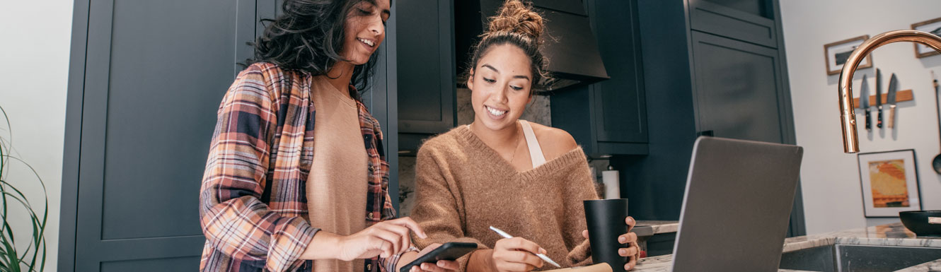 Two young women are in a kitchen smiling while using a cellphone and laptop.