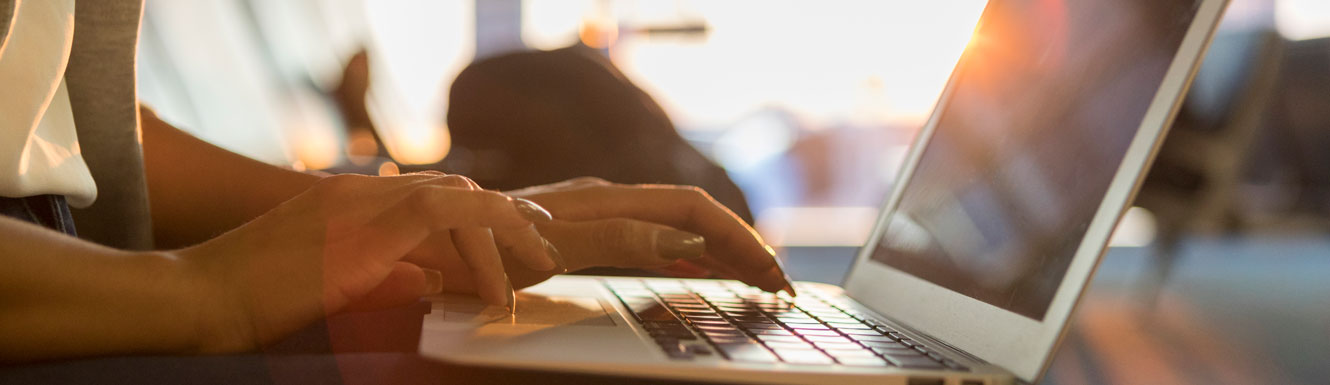 A woman is using a laptop in what appears to be an airport with a blurry bright background.