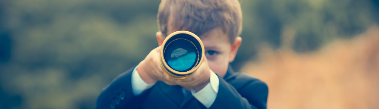 A young boy in a blue business suit is outdoors and looking through a telescope.