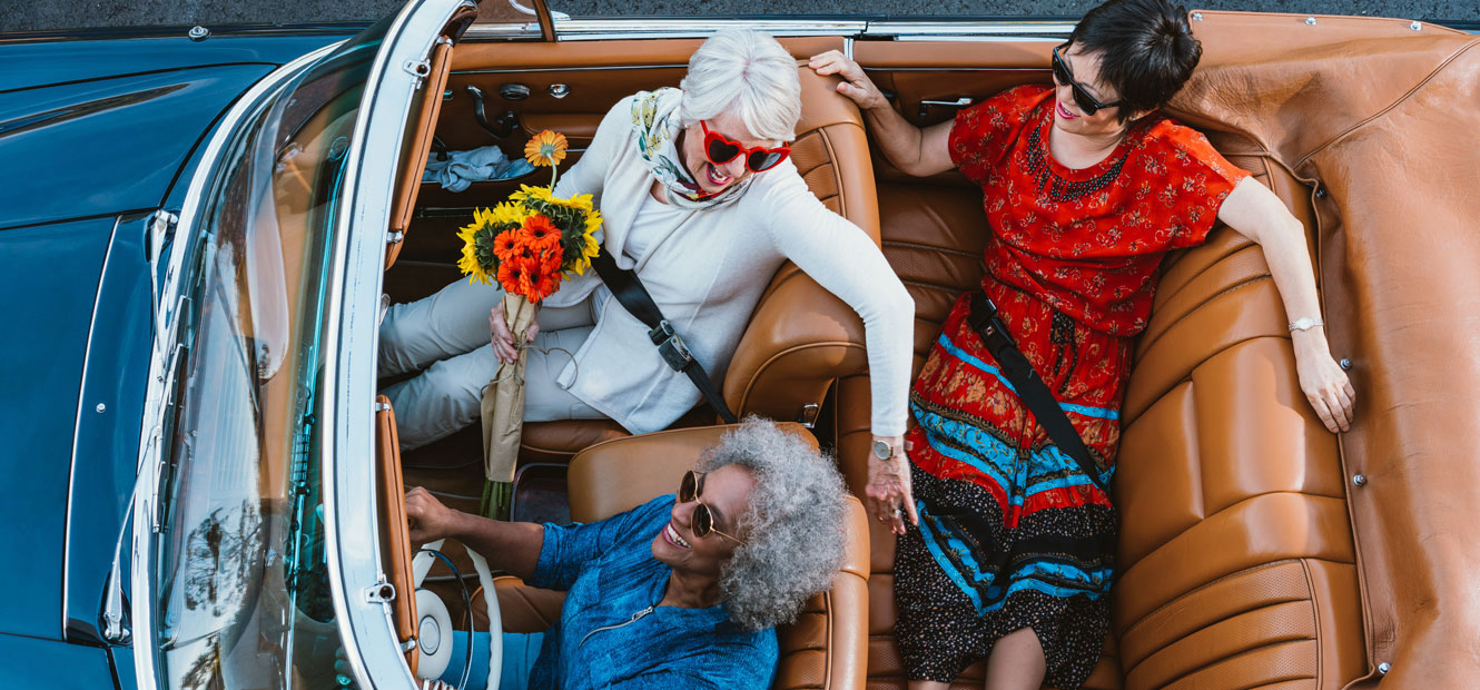 Three friends are shown, all mature women, in a convertible on a road trip.