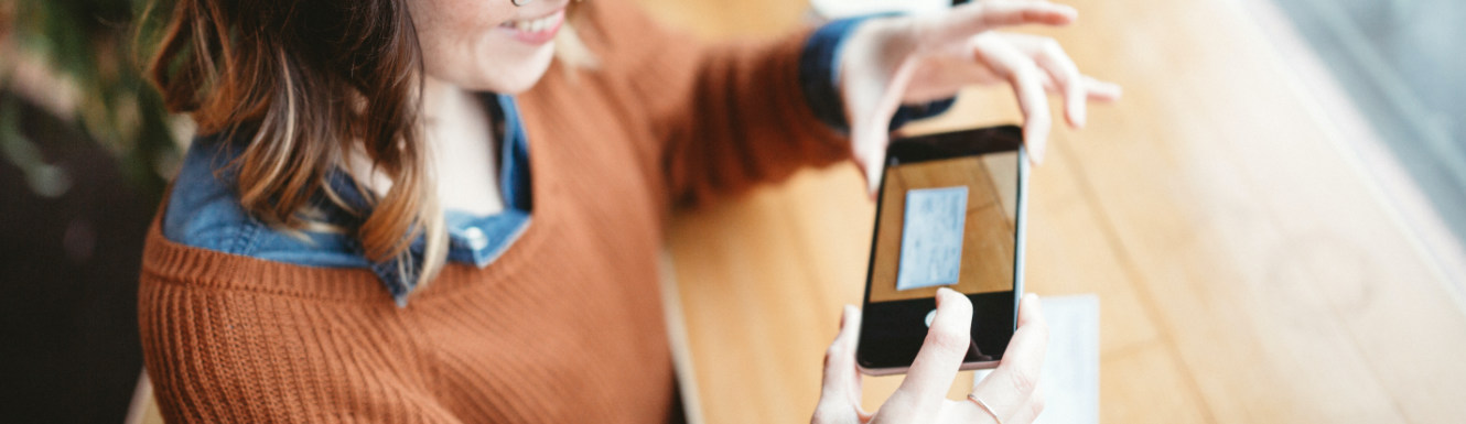 A young woman is sitting at a wooden table and depositing a check using her cellphone.